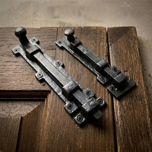 Puede incluir: Dos cerrojos de puerta de metal gris oscuro de estilo antiguo. Cada cerrojo tiene una base rectangular, una barra deslizante y un pomo redondo. Los cerrojos están montados en una puerta de madera con vetas visibles.