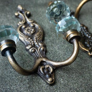 May include: A pair of wall-mounted hooks featuring an ornate, antique bronze-finished metal backplate with intricate scrollwork and a central floral motif, topped with a faceted, clear glass knob at the end of each curved arm.