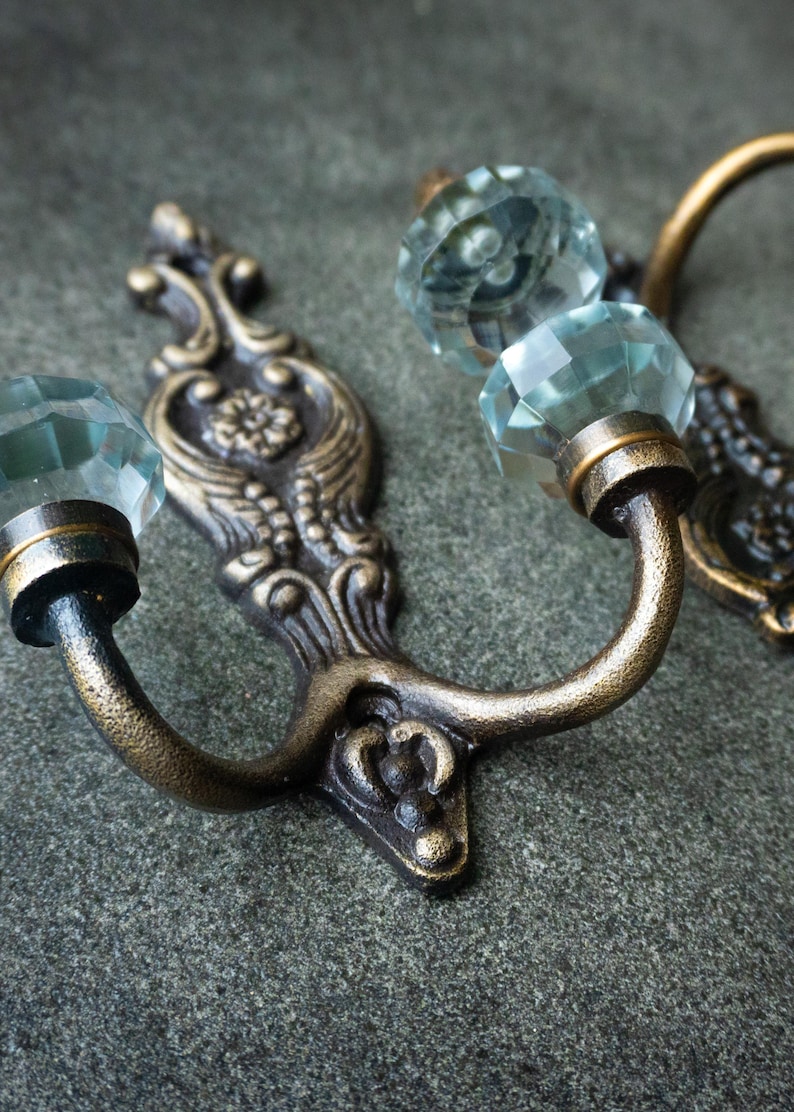 May include: A pair of wall-mounted hooks featuring an ornate, antique bronze-finished metal backplate with intricate scrollwork and a central floral motif, topped with a faceted, clear glass knob at the end of each curved arm.