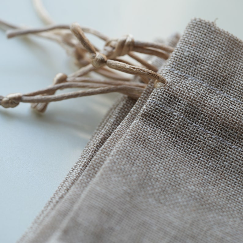 Op de afbeelding: Een close-up van een beige linnen trekkoordtas met een geknoopte sluiting. De tas is gemaakt van natuurlijke stof met een subtiele textuur.