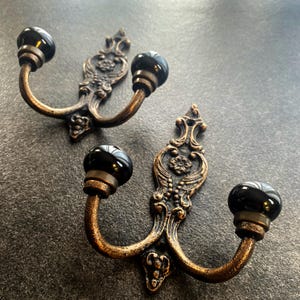 May include: Two antique-style wall hooks with a bronze finish and black, spherical knobs. The hooks have ornate backplates with floral and scroll details, perfect for adding a vintage touch to any space.