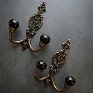 May include: Two ornate, antique-style wall hooks. Each hook features a dark, spherical knob and a decorative, bronze-colored backplate with floral and scrollwork details. The hooks are made of metal and are designed for hanging items.