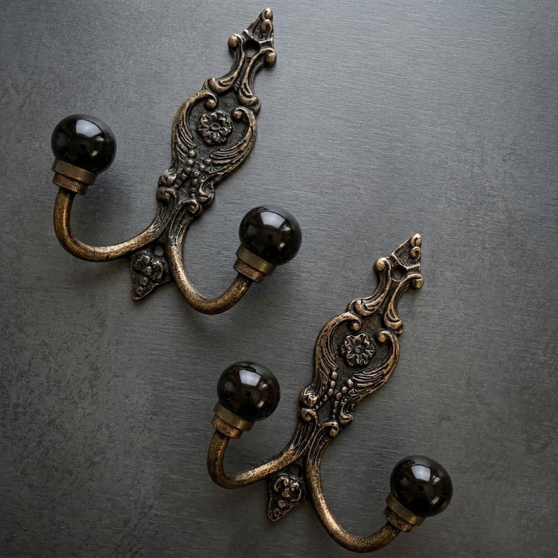 May include: Two ornate, antique-style wall hooks. Each hook features a dark, spherical knob and a decorative, bronze-colored backplate with floral and scrollwork details. The hooks are made of metal and are designed for hanging items.