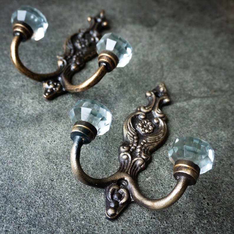 May include: Two antique-style wall hooks with a bronze finish. Each hook features a decorative backplate and two curved arms, each topped with a faceted, clear crystal knob. The hooks are designed for hanging items.