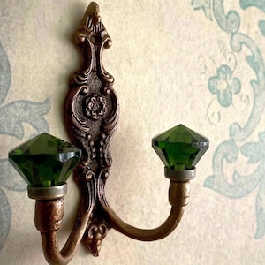 May include: An ornate, bronze-colored wall hook with two curved arms, each holding a faceted, emerald-green glass gem. The hook has a decorative, vintage design with floral and scroll details, mounted on a patterned wall.