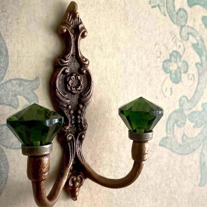 May include: An ornate, bronze-colored wall hook with two curved arms, each holding a faceted, emerald-green glass gem. The hook has a decorative, vintage design with floral and scroll details, mounted on a patterned wall.