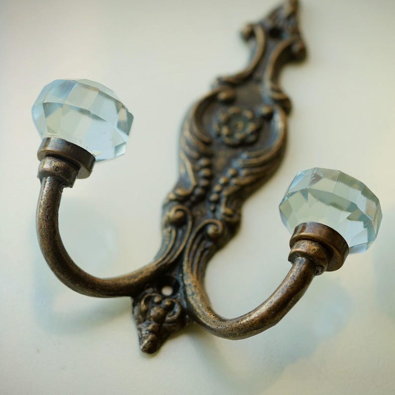 May include: An antique-style double hook with a bronze finish. The hook features two curved arms, each topped with a faceted, light blue crystal knob. The decorative backplate has ornate floral and scroll detailing.