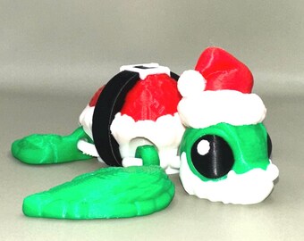 May include: A 3D-printed Christmas turtle figurine. The turtle has a green body, a red Santa hat with white trim, and a black and white shell. The turtle's head has a large black eye. The figurine is designed to be festive and decorative.