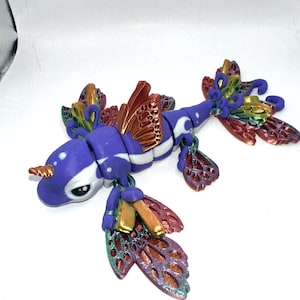 May include: A whimsical, handcrafted sculpture of a narwhal-dragon hybrid. The creature is primarily purple with white accents, a golden horn, and iridescent wings in shades of red, gold, and green. The sculpture has a detailed, fantasy-inspired design.