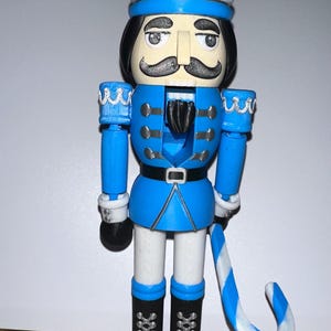 May include: A blue and white decorative nutcracker figurine. The figurine has a blue jacket, white pants, and black boots. It has a black mustache and a blue and white hat. It is holding a blue and white striped cane.