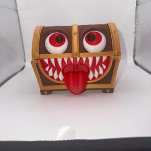 May include: A brown and gold treasure chest with large red and white eyes, a toothy grin, and a red tongue. The chest is 3D printed and has a cartoonish, monster-like appearance.
