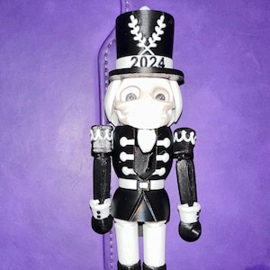 May include: A black and white nutcracker figurine with a top hat that reads "2024". The figurine has a white face with a mask, a black uniform, and black boots with skull and crossbones. The figurine is standing on a purple surface.