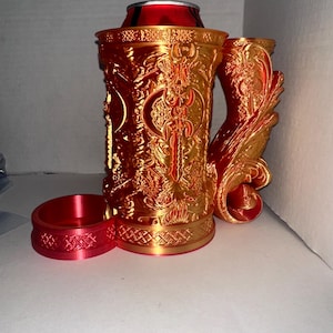 May include: A decorative red and gold beer stein with intricate embossed detailing. The stein features a handle and a separate coaster. A red can is inside the stein. The design includes an axe and other ornate patterns.
