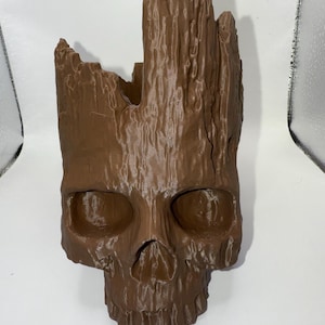 May include: A brown, skull-shaped planter with a textured, wood-like appearance. The planter features large eye sockets and a nose cavity, with the top portion resembling a broken tree trunk. The overall design is a unique blend of nature and macabre.