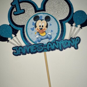May include: A cake topper with a Mickey Mouse theme, featuring the number 1, Mickey Mouse ears, and balloons. The topper is blue and silver glitter with the name "JAMES ANTONY". It is attached to a wooden stick for easy placement.