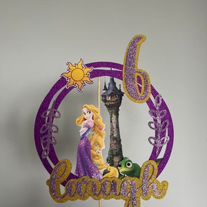 May include: Purple and gold glitter cake topper featuring a cartoon image of Rapunzel, a tower, a chameleon, and the sun. The topper also includes the number 6 and the name "Rapunzel".