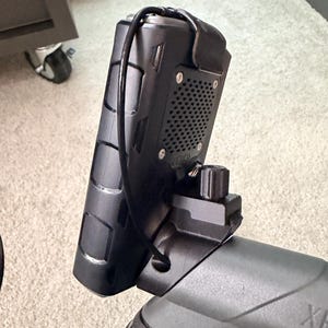 Könnte beinhalten: Ein schwarzes elektronisches Gerät mit strukturierter Oberfläche, montiert an einem grauen Lenker. Das Gerät hat ein Lautsprechergitter und ist mit einem schwarzen Kabel befestigt. Ein schwarzer Knopf ist sichtbar. Der Hintergrund ist ein heller Teppich.