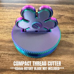 May include: A compact thread cutter in a flower shape, with a circular base and a multi-colored design. The text "COMPACT THREAD CUTTER" and "***45mm ROTARY BLADE NOT INCLUDED***" are printed on the base.