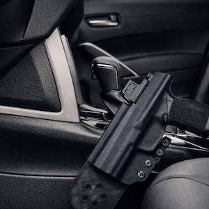 May include: Black handgun in a black holster, mounted inside a car. The holster is attached to the car's interior, near the center console. The image is in black and white, highlighting the gun and holster.