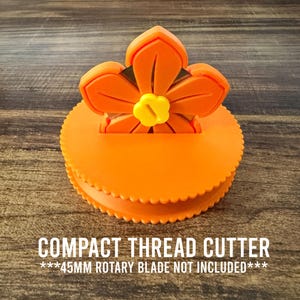 May include: A bright orange circular thread cutter featuring a flower-shaped top with a small yellow center knob. The device includes the text COMPACT THREAD CUTTER and ***45MM ROTARY BLADE NOT INCLUDED***. The base has a scalloped edge design.