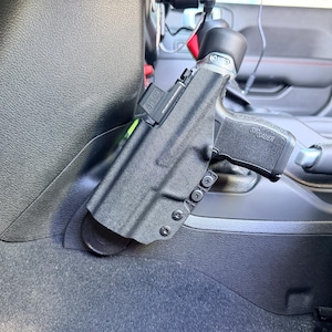 May include: Black handgun in a black holster mounted inside a vehicle. The handgun is a Sig Sauer model. The holster is attached to the vehicle's interior. The vehicle's interior is black and green.