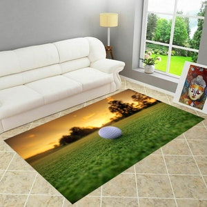 May include: A golf ball sits on a green putting green with a sunset in the background. The green is lush and the sunset is a warm orange and yellow.