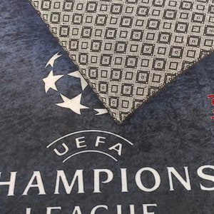 Puede incluir: Una alfombra de puerta de tela azul y negra con el logotipo y el texto de la UEFA Champions League. La alfombra tiene una superficie texturizada y una etiqueta de muestra.