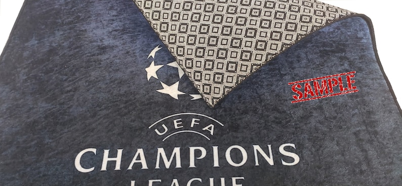 Puede incluir: Una alfombra de puerta de tela azul y negra con el logotipo y el texto de la UEFA Champions League. La alfombra tiene una superficie texturizada y una etiqueta de muestra.
