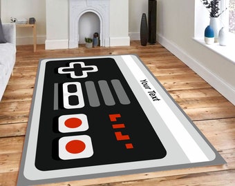 Puede incluir: Una alfombra gris y negra con un diseño de mando de videojuego retro. El mando tiene un signo más blanco, botones grises y círculos rojos. El texto "Your Text" está impreso en la alfombra.