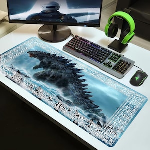 Può includere: Un tappetino per mouse rettangolare a tema Godzilla con un'illustrazione dettagliata del mostro che emerge dall'oceano. Il tappetino ha un bordo decorativo blu e bianco, con tastiera, mouse e cuffie.