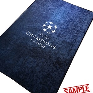 Puede incluir: Una alfombra azul con un logotipo blanco de la UEFA Champions League. El logotipo presenta un bal&oacute;n de f&uacute;tbol con estrellas y el texto "UEFA CHAMPIONS LEAGUE". La alfombra tiene una superficie texturizada y est&aacute; marcada con "SAMPLE".