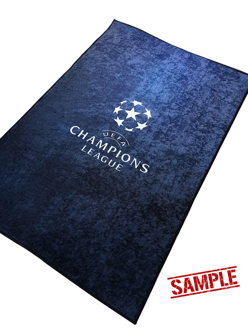 Puede incluir: Una alfombra azul con un logotipo blanco de la UEFA Champions League. El logotipo presenta un bal&oacute;n de f&uacute;tbol con estrellas y el texto "UEFA CHAMPIONS LEAGUE". La alfombra tiene una superficie texturizada y est&aacute; marcada con "SAMPLE".
