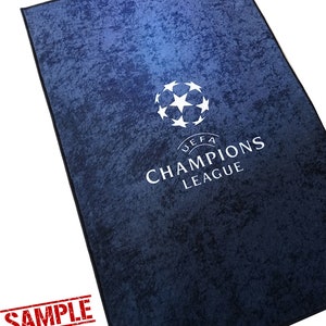 Puede incluir: Una alfombra azul y blanca de la Liga de Campeones con el logotipo y el texto de la UEFA Champions League. La alfombra tiene un aspecto desgastado y texturizado.