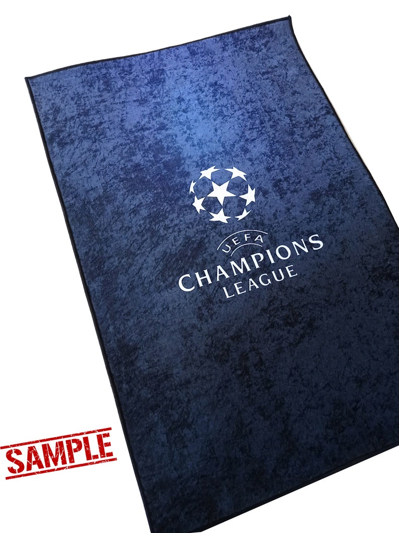 Puede incluir: Una alfombra azul y blanca de la Liga de Campeones con el logotipo y el texto de la UEFA Champions League. La alfombra tiene un aspecto desgastado y texturizado.