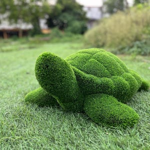 Könnte beinhalten: Eine dekorative Schildkrötenfigur, die mit leuchtend grünem Kunstrasen bedeckt ist. Die Skulptur weist eine moosartige, texturierte Oberfläche mit einem detaillierten Panzer-Muster, einem abgerundeten Kopf und vier Flossen auf, die eine Meeresschildkröte darstellen.