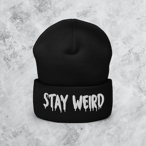 Op de afbeelding: Zwarte beanie met een witte "STAY WEIRD" inscriptie in een druppelend lettertype.