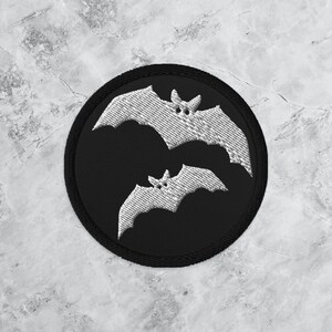 May include: Embroidered patch featuring two white bats on a black circular background. The bats have detailed wing patterns and small eyes. The patch has a black border and is set against a marbled surface.