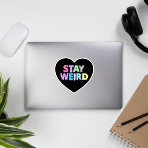 May include: A black heart-shaped sticker with the text "STAY WEIRD" in rainbow colors. The text appears to be dripping, giving it a fun and quirky look.