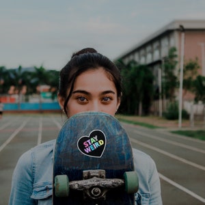 May include: A young person is holding a skateboard in front of their face. The skateboard has a black heart sticker with the words "Stay Weird" in rainbow colors.