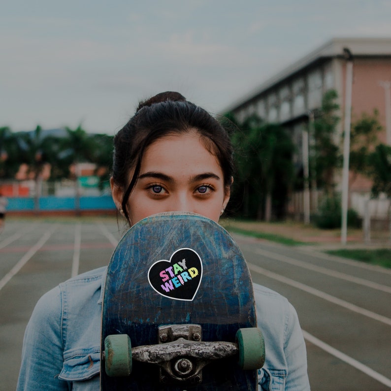 May include: A young person is holding a skateboard in front of their face. The skateboard has a black heart sticker with the words "Stay Weird" in rainbow colors.
