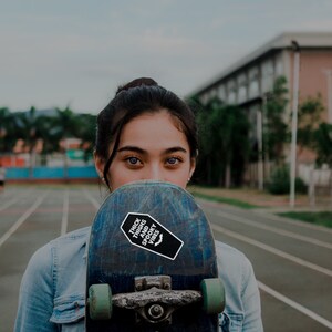 May include: A young person with dark hair is holding a skateboard in front of their face. The skateboard has a black and white sticker that says "Thick Thighs and Spooky Vibes" with a bat graphic.