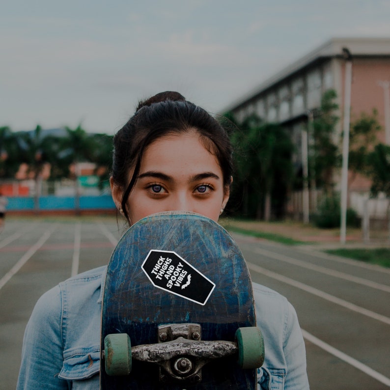 May include: A young person with dark hair is holding a skateboard in front of their face. The skateboard has a black and white sticker that says "Thick Thighs and Spooky Vibes" with a bat graphic.