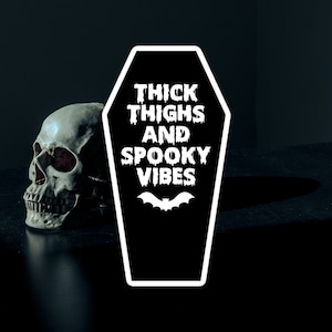 May include: A black and white coffin-shaped sticker with the text "Thick Thighs and Spooky Vibes" and a bat graphic. The sticker is on a dark surface next to a white skull.