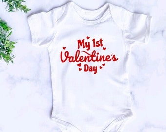 May include: White baby onesie with red text that says "My 1st Valentine's Day" with small red hearts around the text.