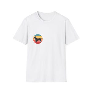 Pode incluir: Camiseta branca com um gráfico circular retrô de um cão dachshund preto em um fundo de listras horizontais vermelhas, laranja, amarelas e azuis. A camiseta tem mangas curtas e gola redonda.