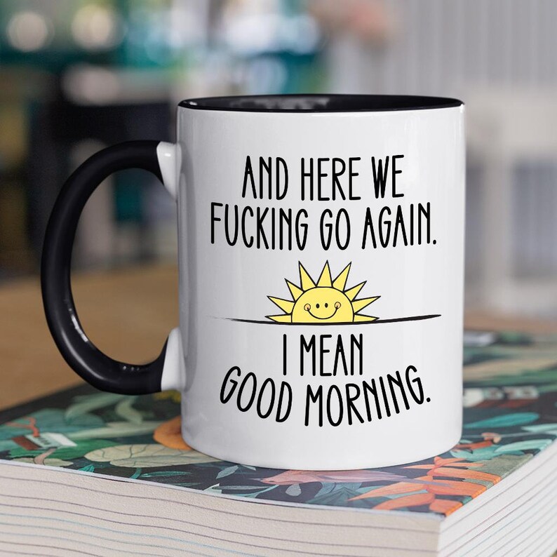 Pode incluir: Caneca de caf&eacute; branca com al&ccedil;a preta e borda preta. A caneca tem um ditado engra&ccedil;ado que diz "And here we fucking go again. I mean good morning." com um sol de desenho animado com uma cara sorridente.