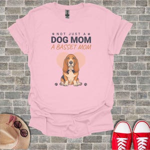 Pode incluir: Uma t-shirt de manga curta rosa claro com um gráfico de um cão Basset Hound castanho e branco centrado à frente de um coração rosa claro. O texto diz NOT JUST A DOG MOM A BASSET MOM em letras cinzento-escuras e laranja com pegadas.