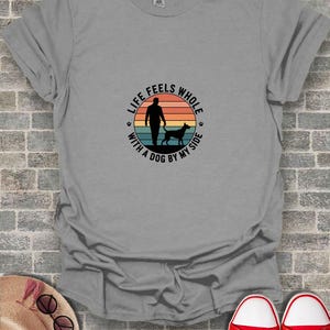 Pode incluir: Camiseta cinza com um gráfico circular com o texto "LIFE FEELS WHOLE WITH A DOG BY MY SIDE". O gráfico mostra uma pessoa e um cão contra um pôr do sol. Tênis vermelhos visíveis na parte inferior.