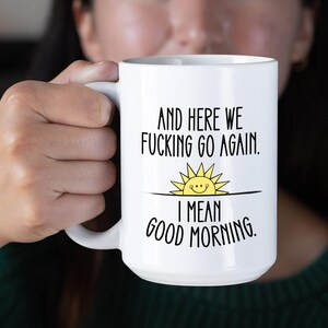 Puede incluir: Taza de caf&eacute; de cer&aacute;mica blanca con texto negro que dice "And here we fucking go again. I mean good morning." Un sol amarillo de dibujos animados con una cara sonriente est&aacute; sobre el texto.