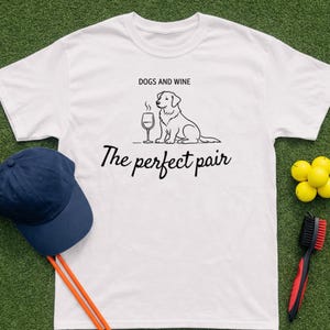 Pode incluir: Camiseta branca com o texto "DOGS AND WINE" acima de um gráfico de um cão e um copo de vinho. Abaixo do gráfico está o texto "The perfect pair". Um boné azul marinho, bolas de golfe e uma escova de taco de golfe também são visíveis.